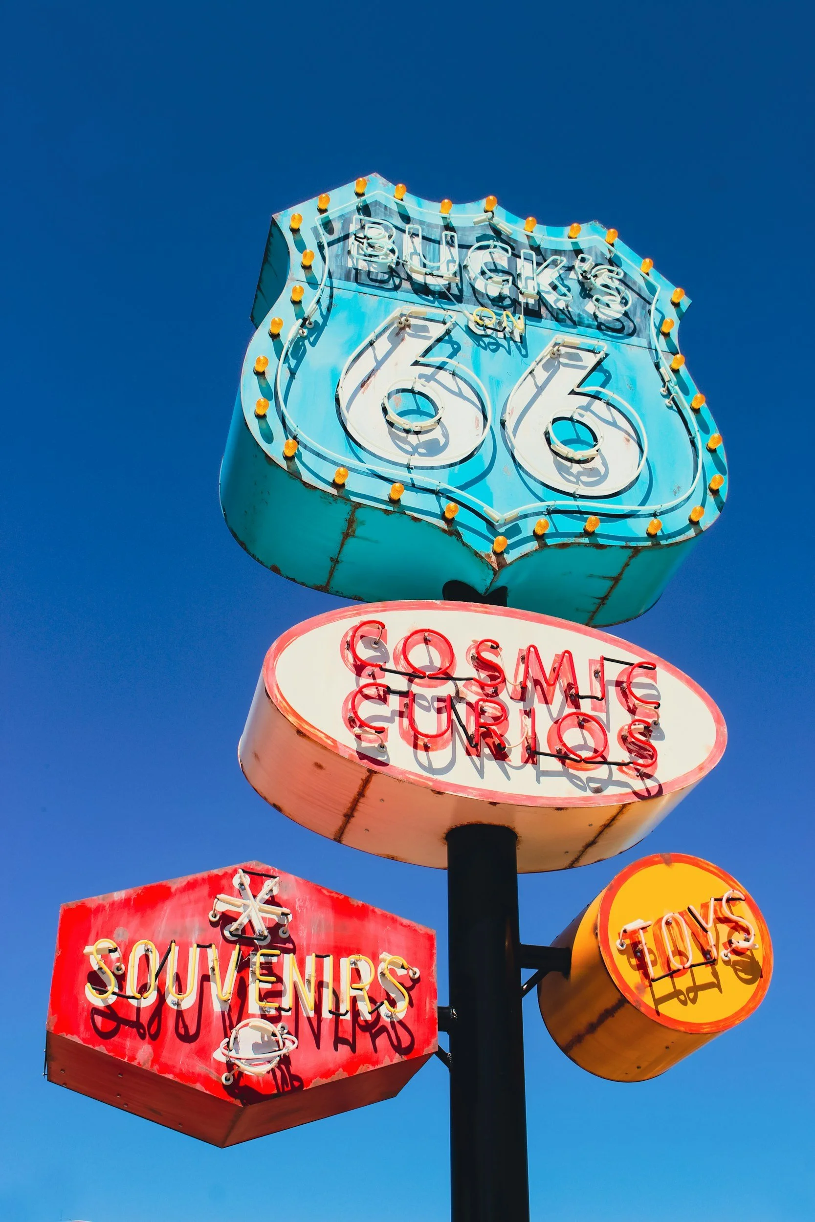 Neon signs advertising Buck's Route 66, souvenirs, toys, and gum, against a clear blue sky.