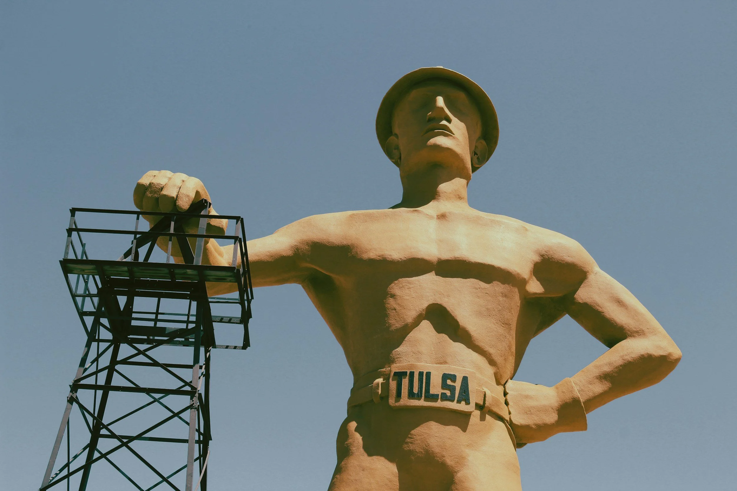 A large statue of a man with a hat, shirtless, with the word 'Tulsa' on his belt, standing with hands on hips, against a clear blue sky, with a metal structure nearby.