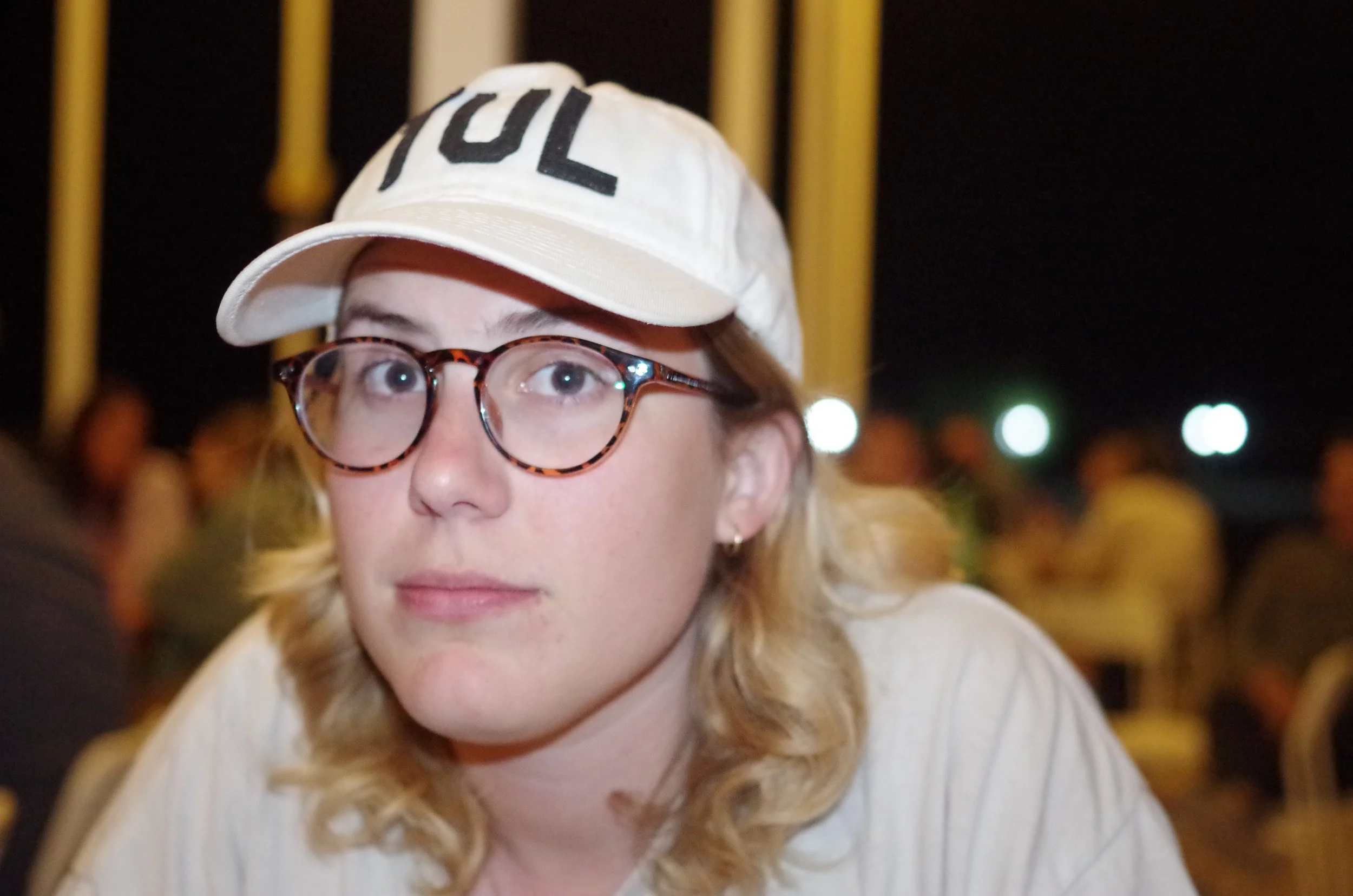 A young woman with curly blonde hair, wearing round tortoiseshell glasses, a white cap with black letters, and a white shirt, sitting at an outdoor event at night with other people in the background.