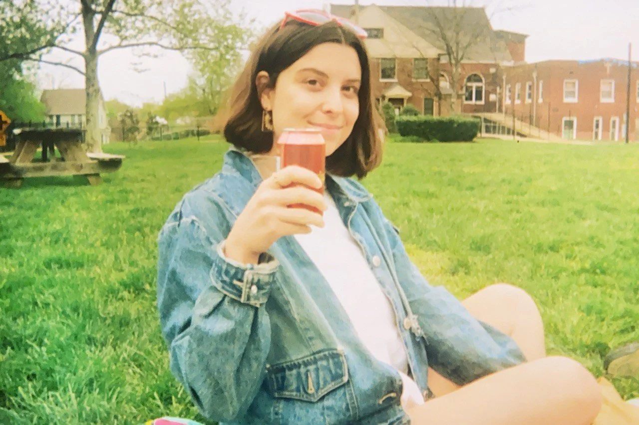 Young woman sitting on grass outdoors holding a can of soda, wearing a denim jacket and sunglasses, with a building and trees in the background.