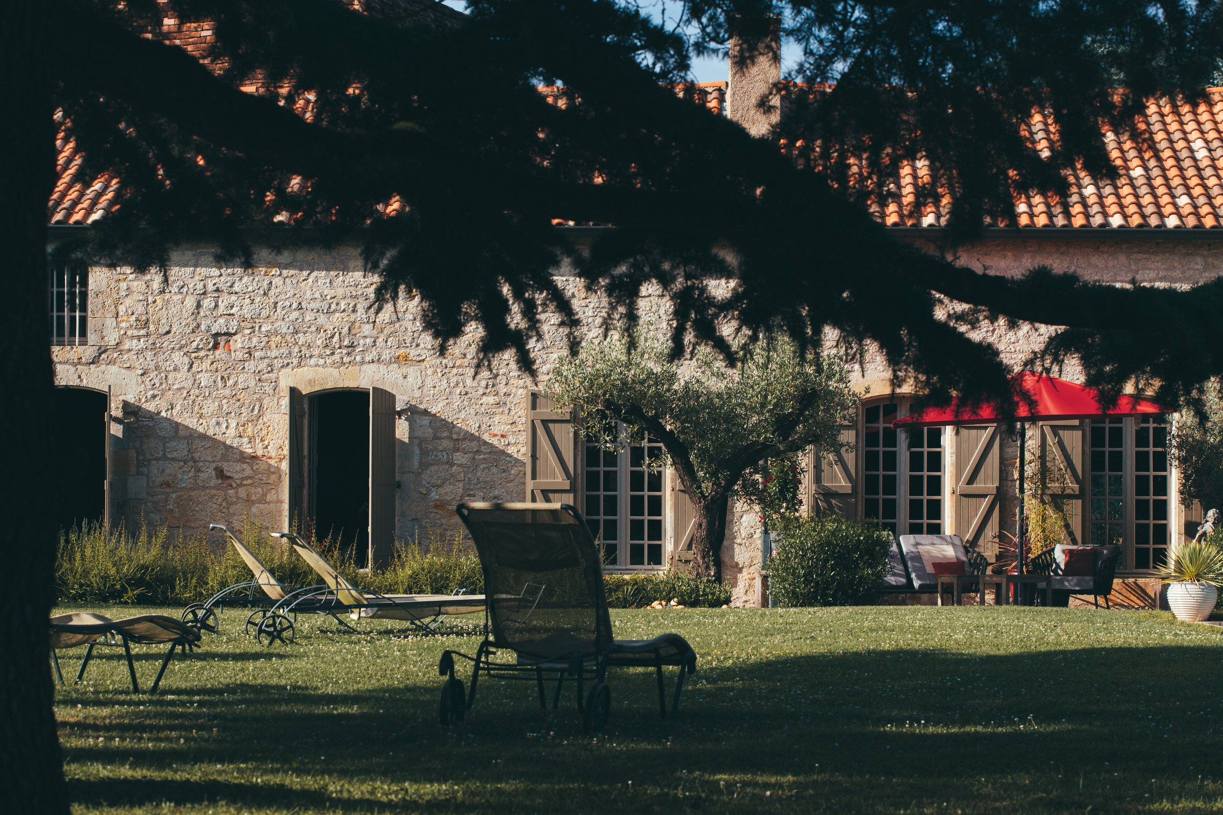 Une maison en pierre avec des fenêtres à volets en bois, un jardin avec des chaises longues et un parasol rouge, des arbres et un mobilier d'extérieur.