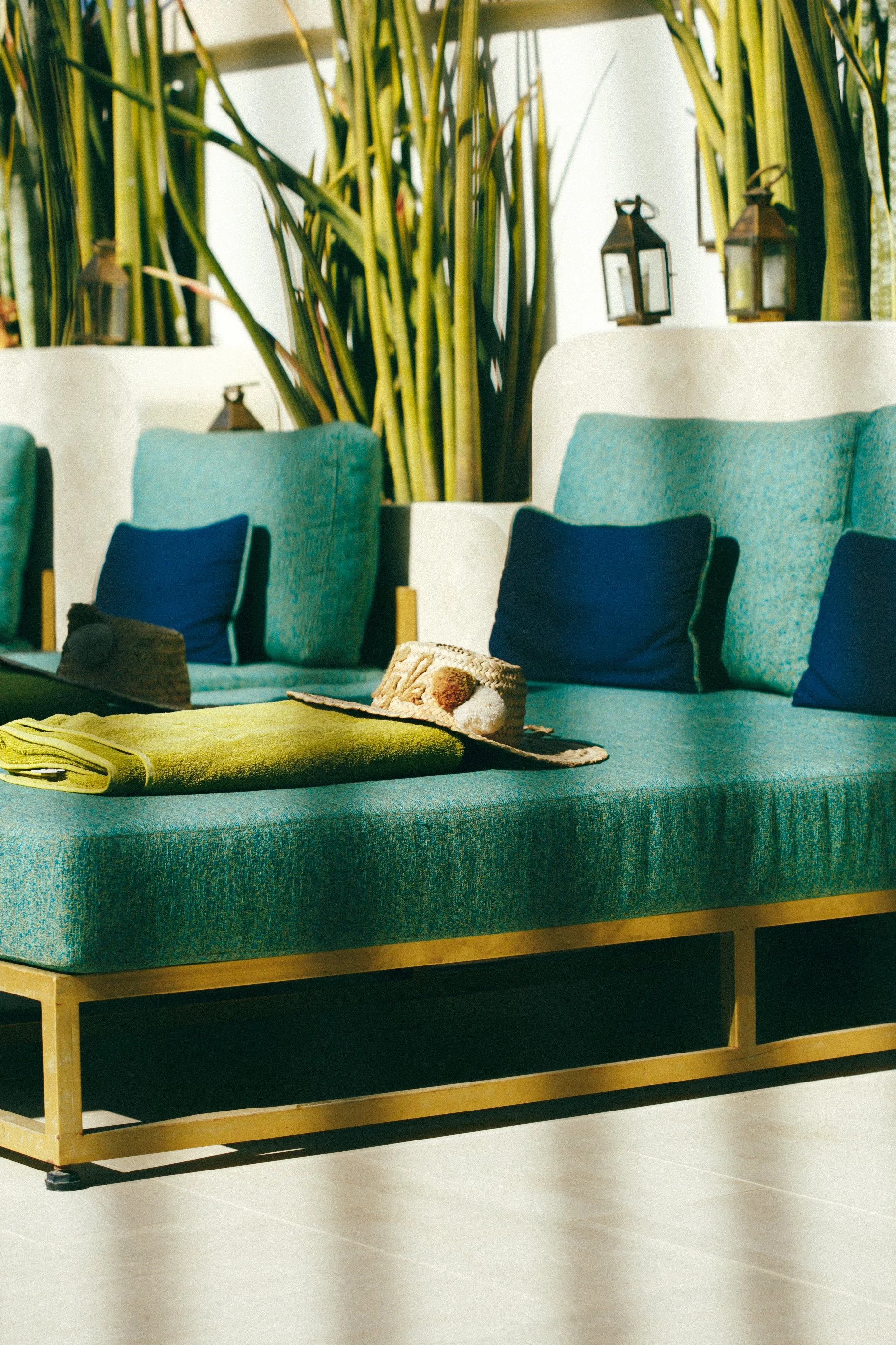 Canapé turquoise avec coussins bleus, toque de soleil et chapeaux posés dessus, dans un salon avec plantes vertes en arrière-plan et lanternes décoratives.