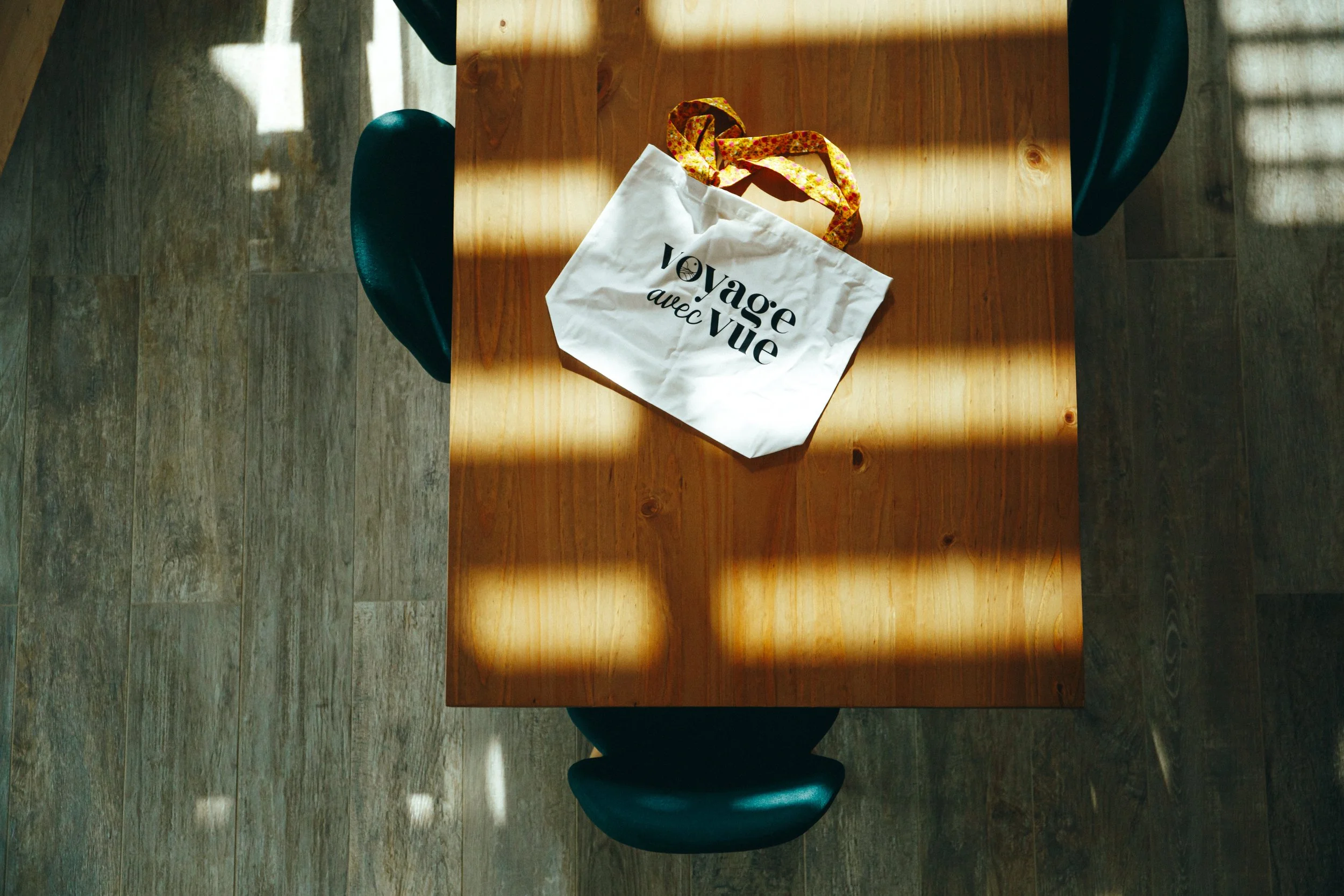 Sac en tissu blanc avec écrit 'voyage avec vue' posé sur une table en bois, éclairée par la lumière naturelle d'une fenêtre.