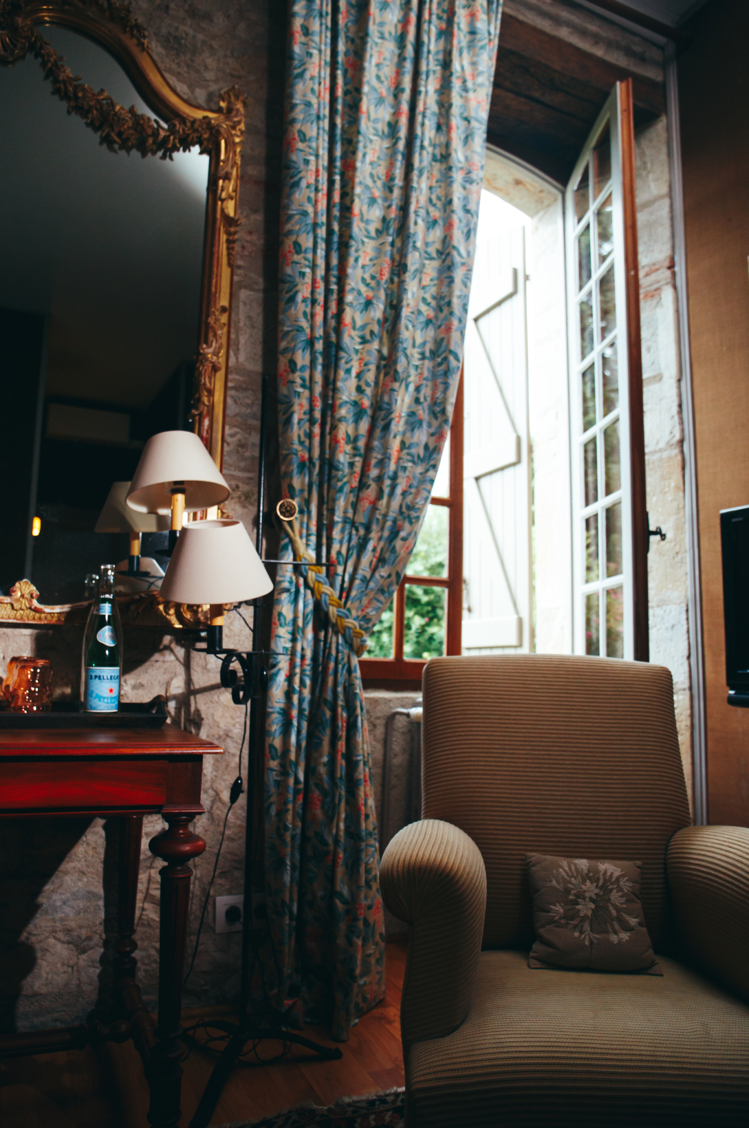 Intérieur d'une pièce avec un fauteuil beige, une lampe à deux abat-jours, une fenêtre ouverte avec des rideaux à motifs floraux, un miroir doré, un petit meuble en bois avec une bouteille d'eau et une lampe, mur en pierre, sol en parquet.