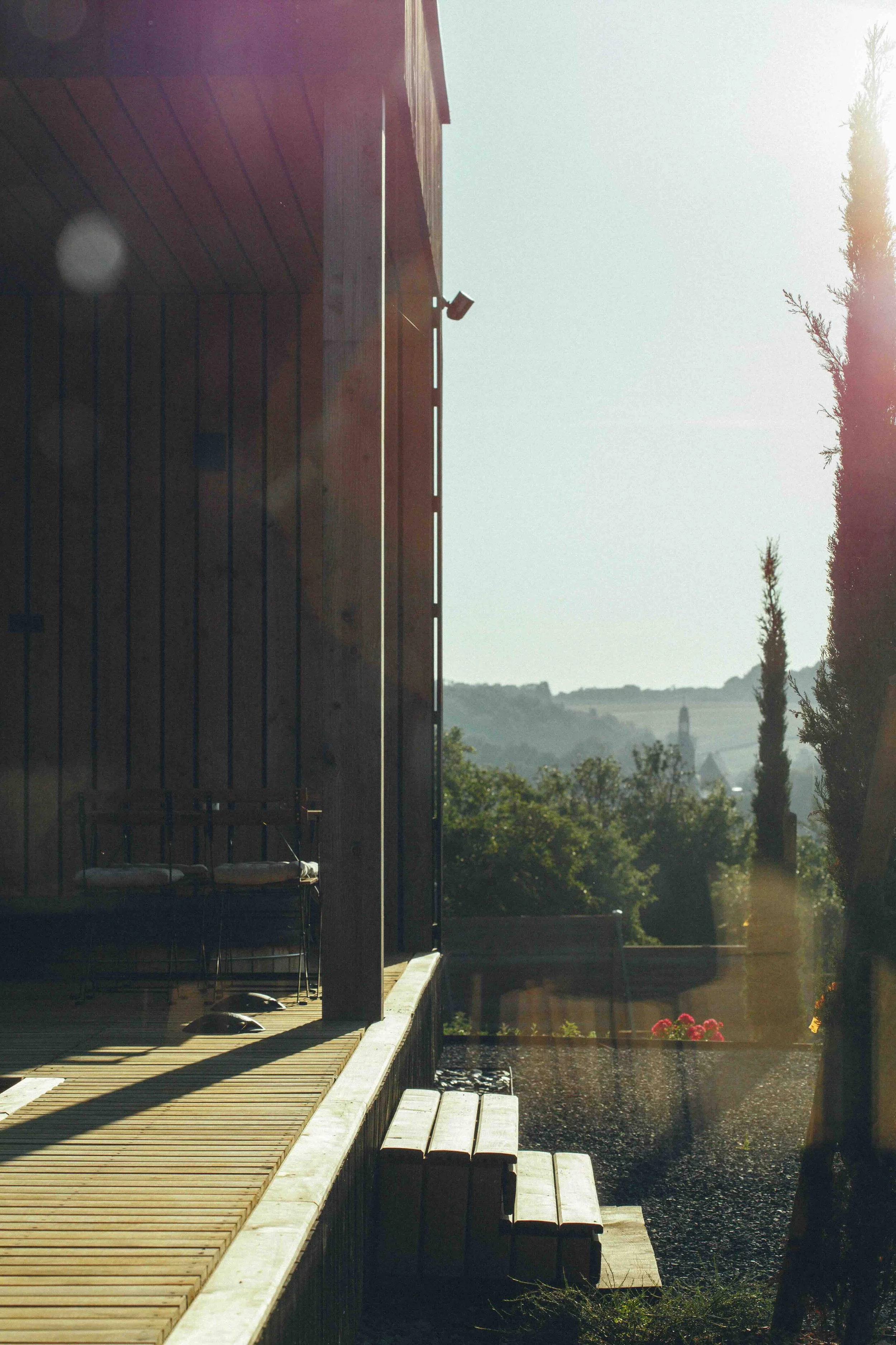 Une vue d'une terrasse en bois attachée à une maison en bois, avec des escaliers en bois, entourée de végétation et avec un paysage de collines en arrière-plan sous un ciel ensoleillé.
