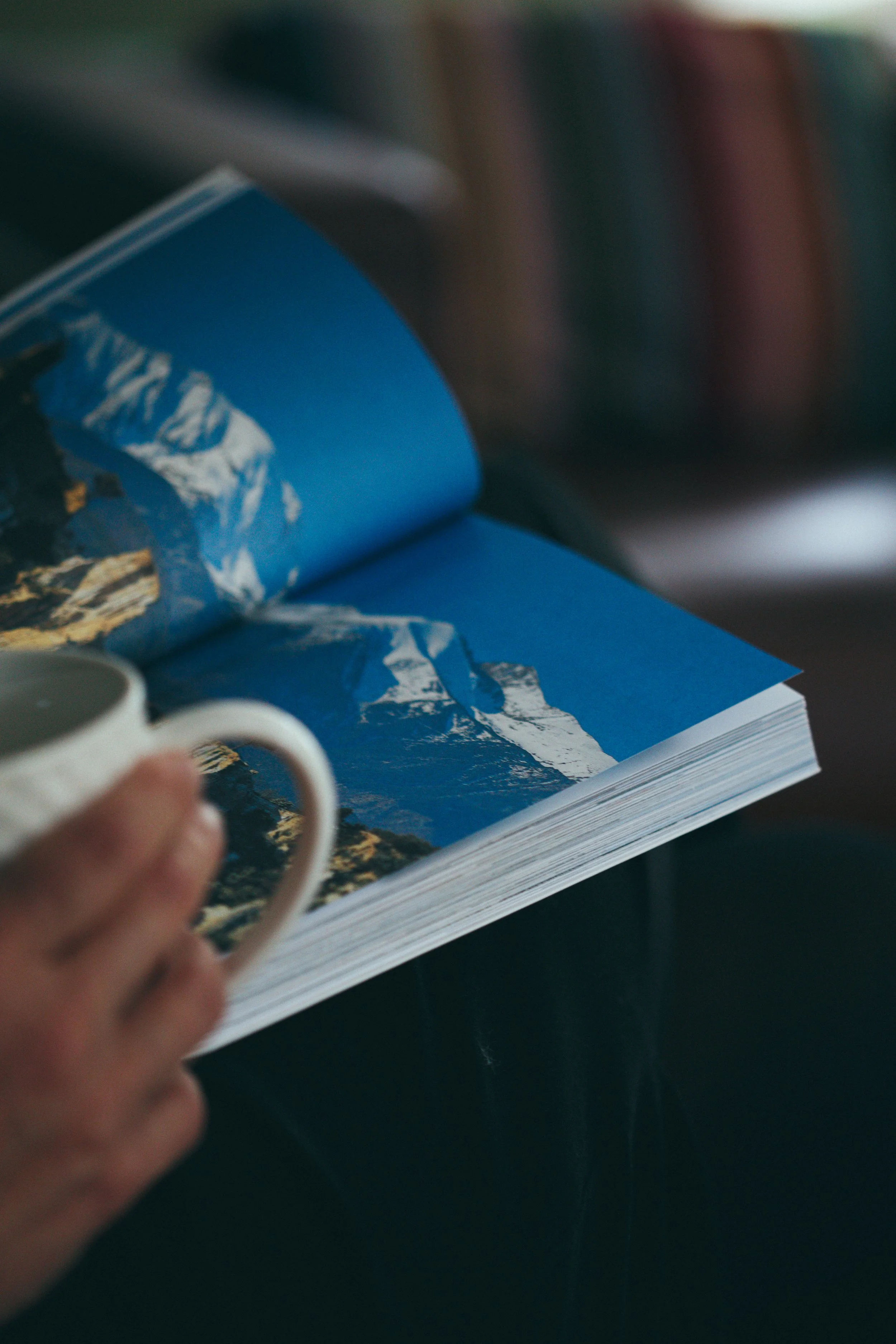 Une main tient une tasse blanche devant un livre ouvert avec une photo de montagnes enneigées sur la page.
