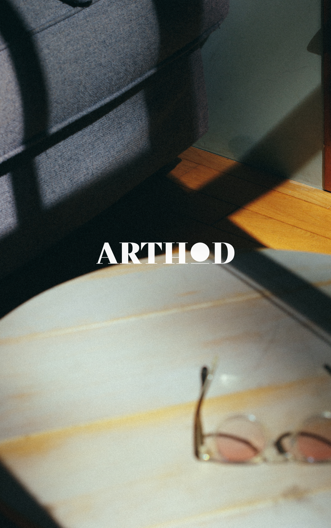 Une lampe posée sur une table en bois, avec des lunettes de soleil à côté, un canapé gris en arrière-plan et une ombre portée sur le mur.