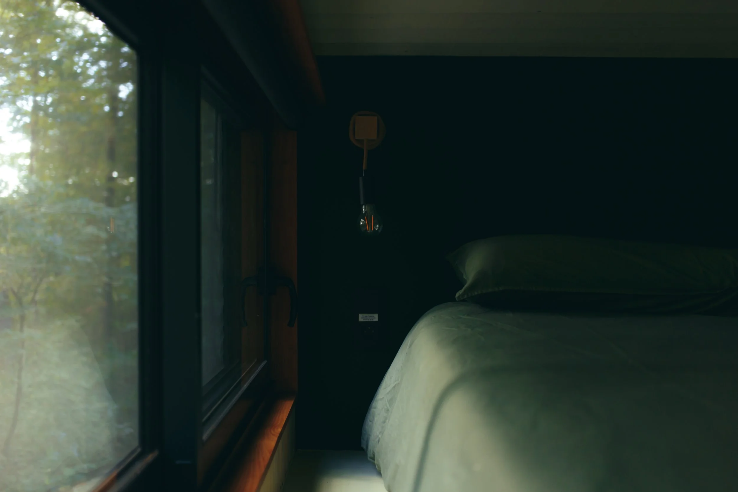 Intérieur sombre d'une chambre avec un lit fait, une lampe suspendue et une fenêtre laissant entrer la lumière naturelle, vue depuis le côté.