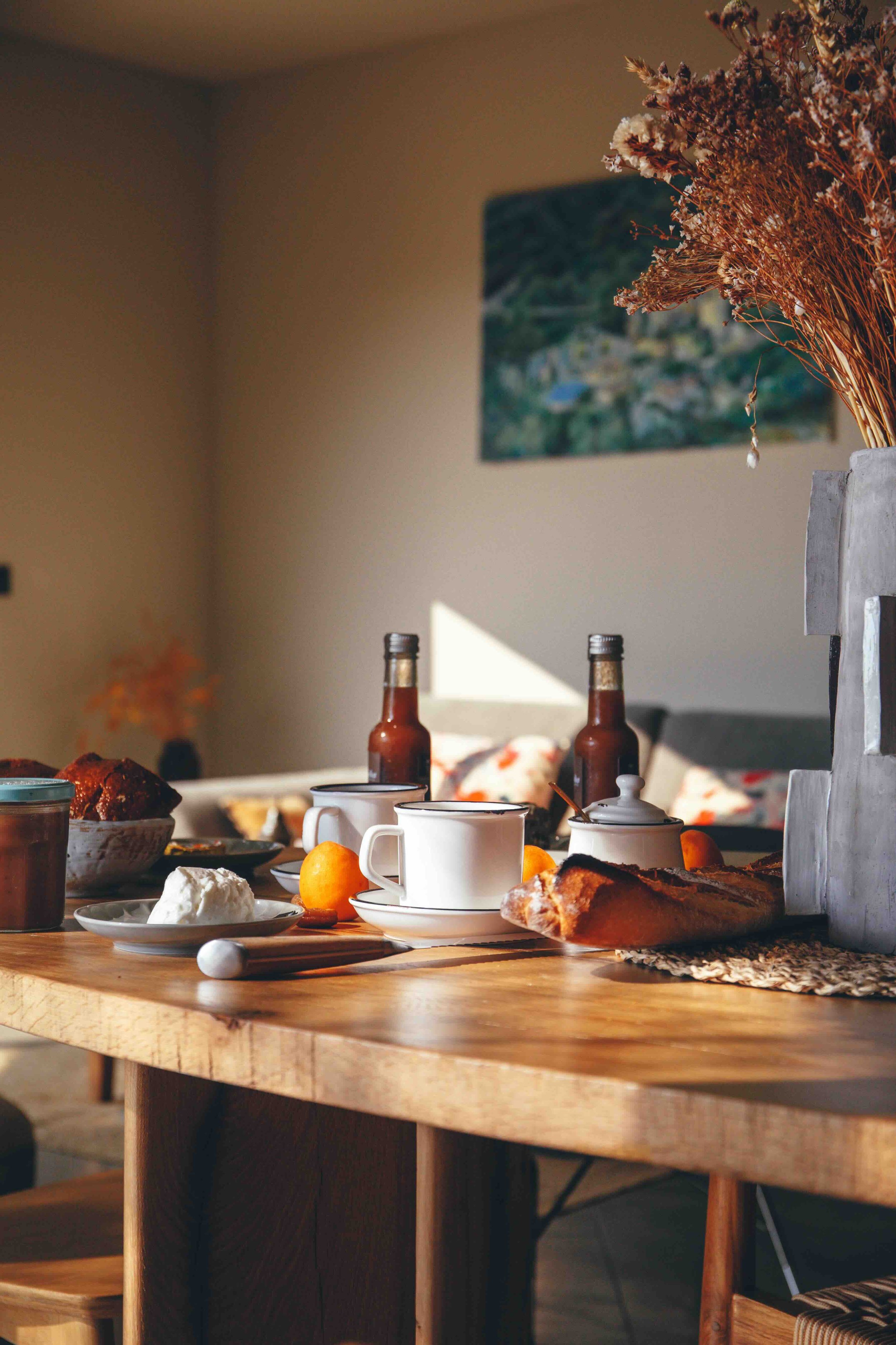 Table de petit déjeuner avec des tasses, une carafe, des fruits, du pain, des œufs, des bouteilles de sauce, dans une pièce illuminée par la lumière naturelle. Décor avec une peinture sur le mur et un vase de fleurs.