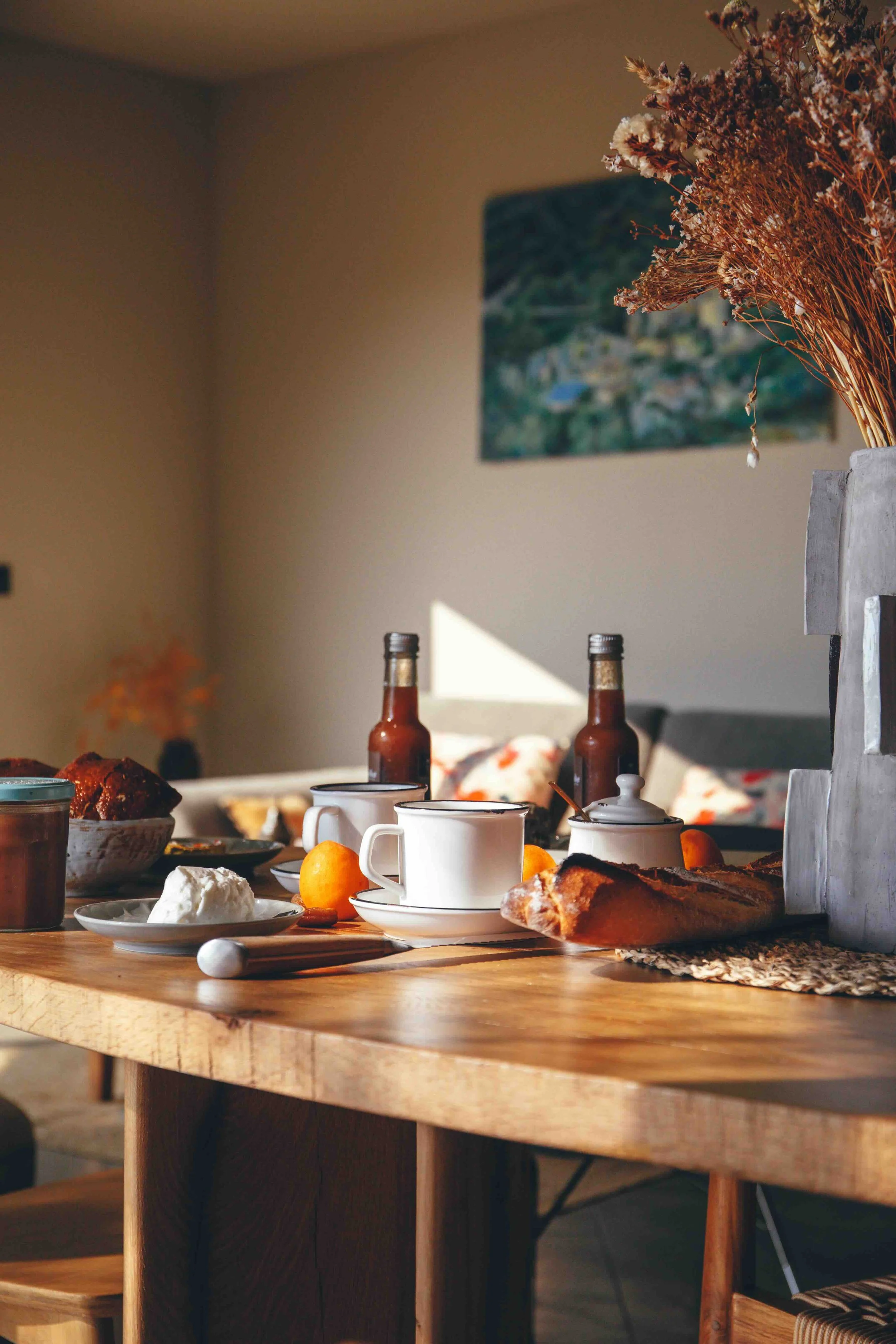 Petit déjeuner avec mugs, bouteilles de sirop, pain, fromage, fruits et confiture sur une table en bois dans une pièce éclairée par la lumière naturelle.