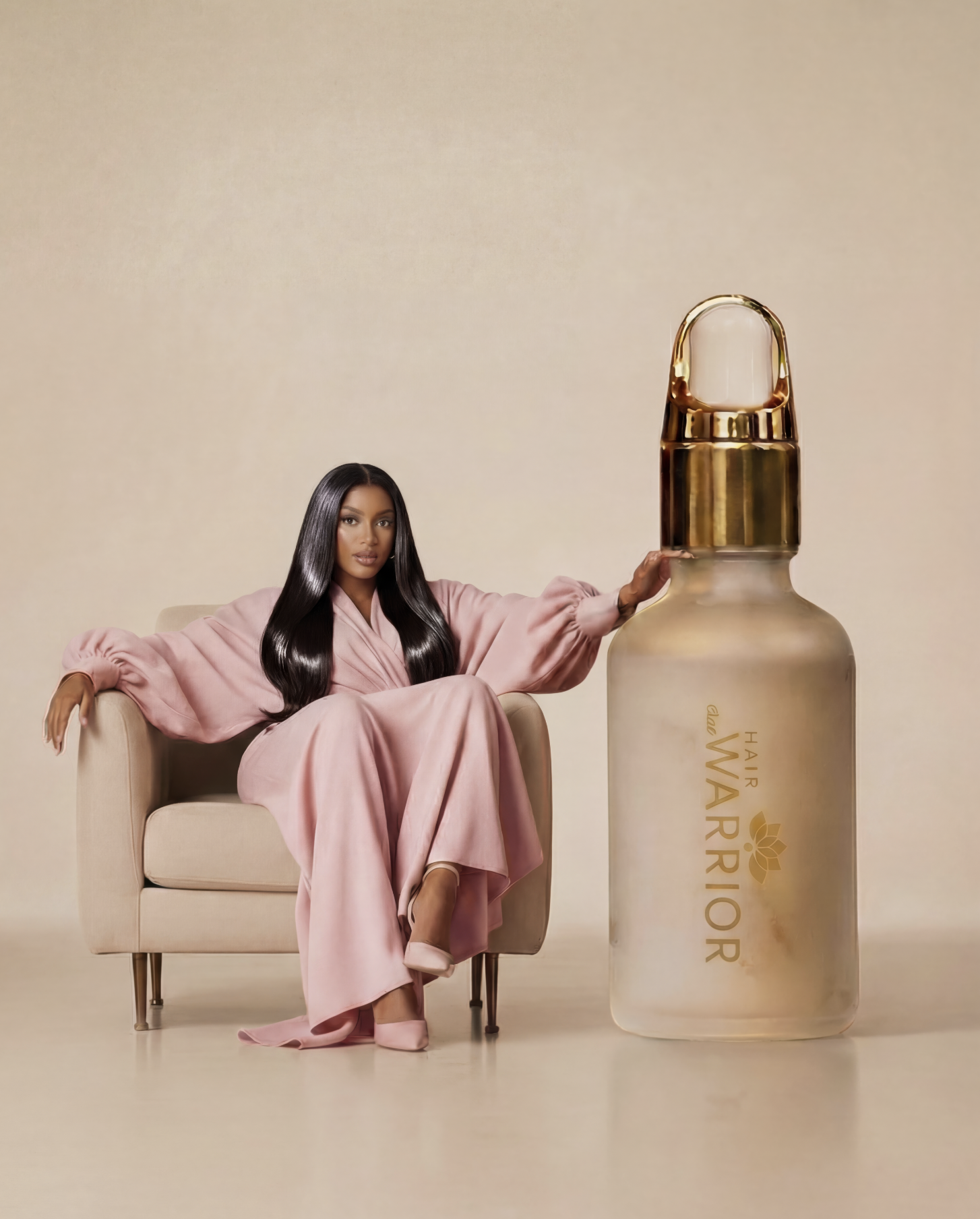 A woman with long dark hair wearing a pink dress sitting on a beige armchair, with her arm resting on a giant bottle of hair oil labeled 'Hair Warrior' and featuring a gold cap and logo.