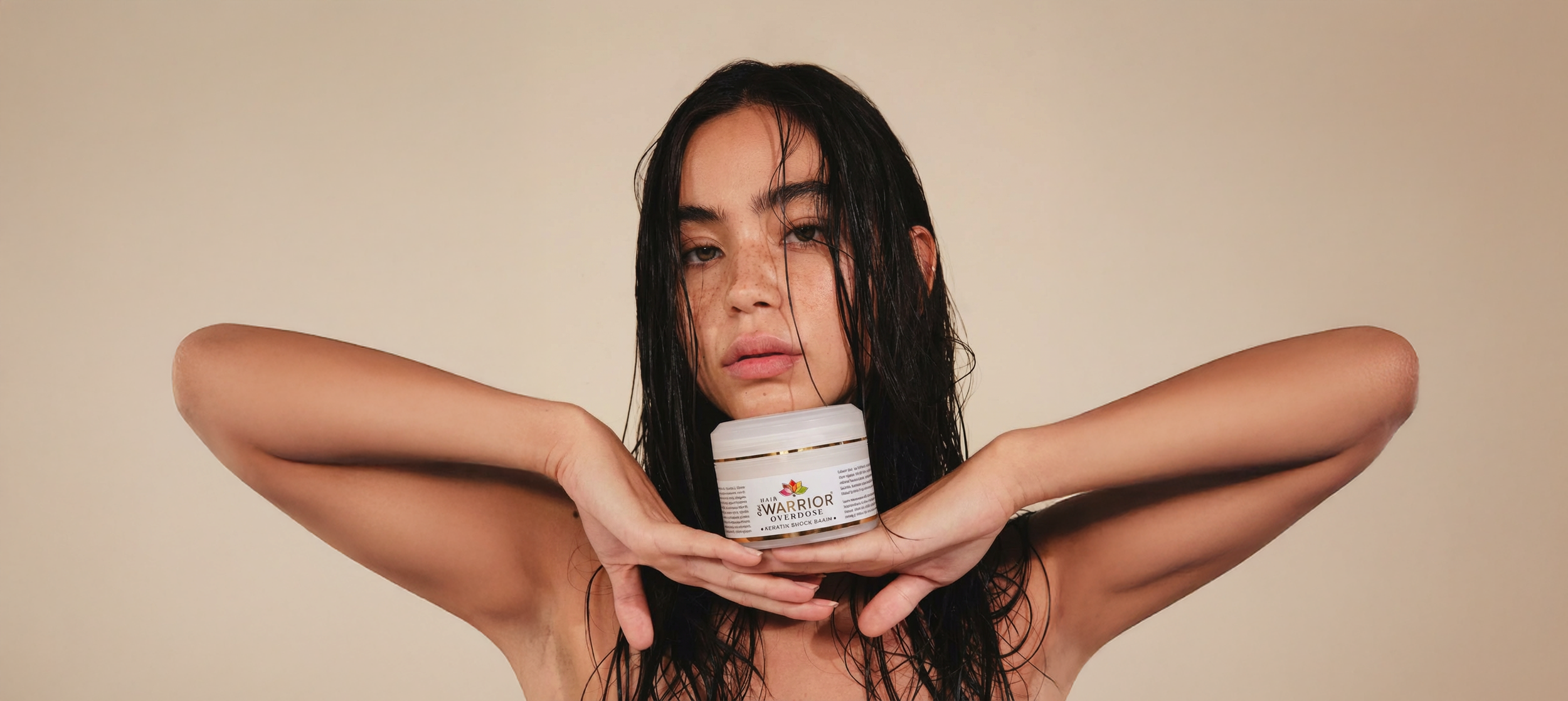 A woman with wet black hair holding a jar of Warrior Overdose hair product under her chin, looking directly at the camera with a neutral expression, against a plain beige background.