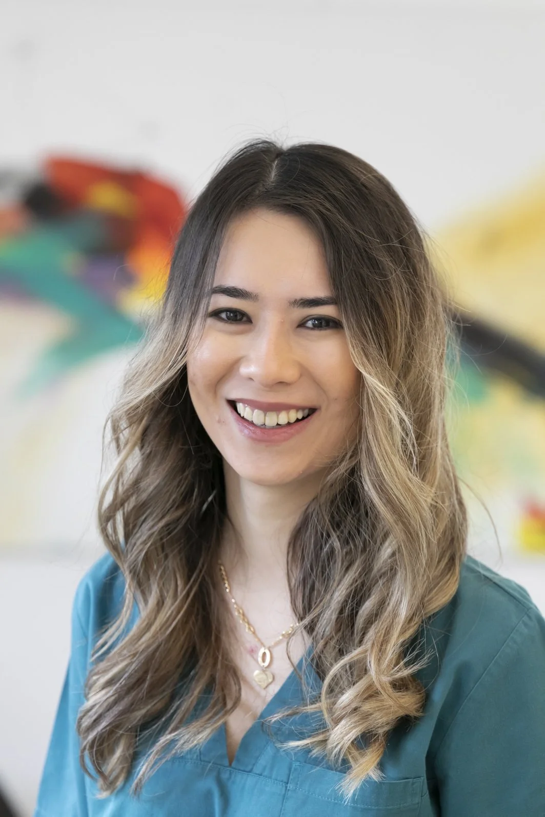 Portrait einer lächelnden Frau mit braunen, lockigen Haaren vor einem bunten, unscharfen Hintergrund.