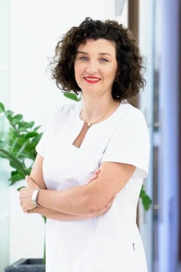 Eine Frau mit braunen, lockigen Haaren trägt ein weißes Kleid und steht in einem modernen, hell erleuchteten Raum mit einer grünen Pflanze im Hintergrund.