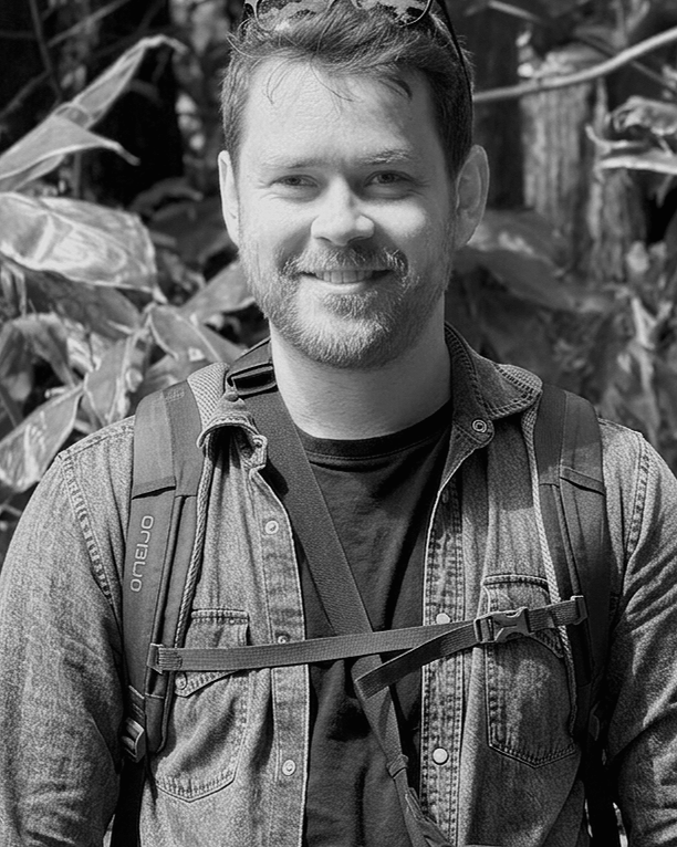 A smiling man with a beard and short hair, wearing a denim jacket and a backpack, standing in front of leafy plants.