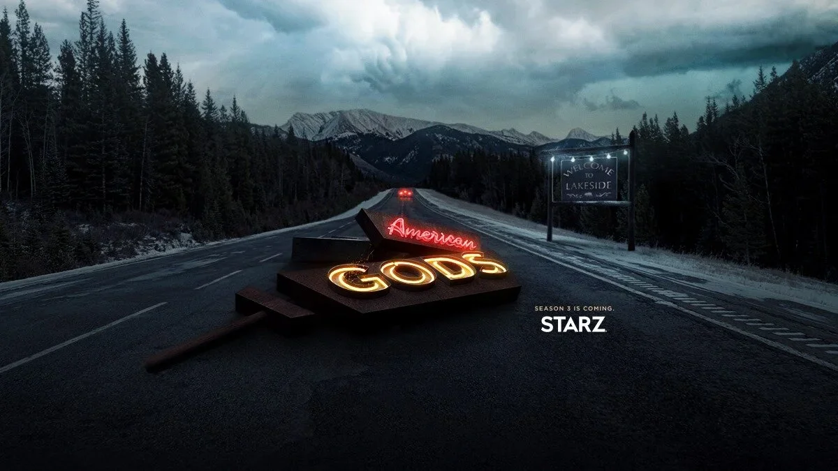 A dark road in a mountainous area with trees on both sides, a sign that reads "Welcome to Lakeside," a neon sign on the road spelling "American Gods," and a small text indicating that season 3 is coming to STARZ.