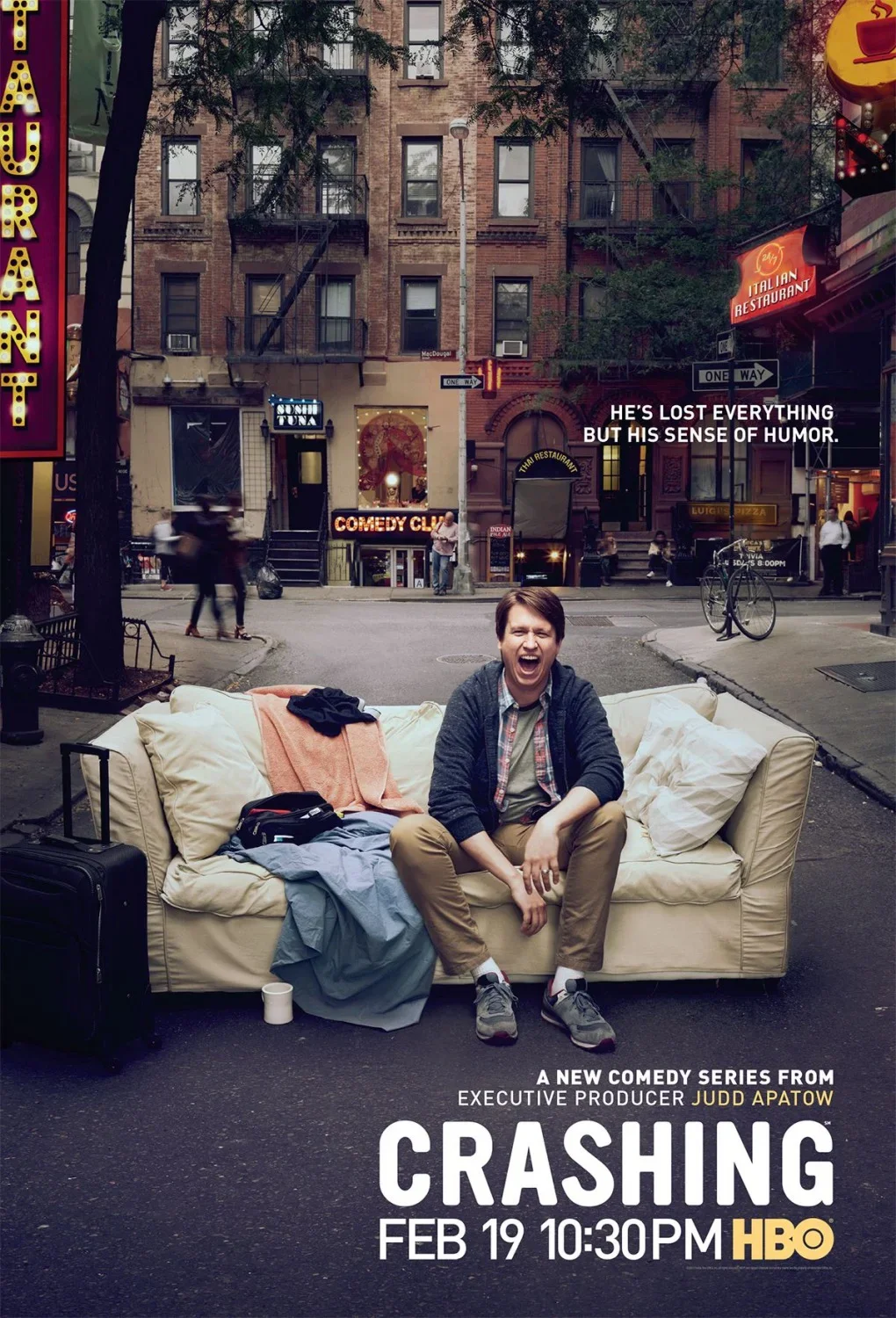 Man sitting on a sofa with a wide smile and laughing, on a city street with storefronts and signs, piles of clothes and belongings around him, advertising a new comedy series called 'Crashing' premiering on HBO.