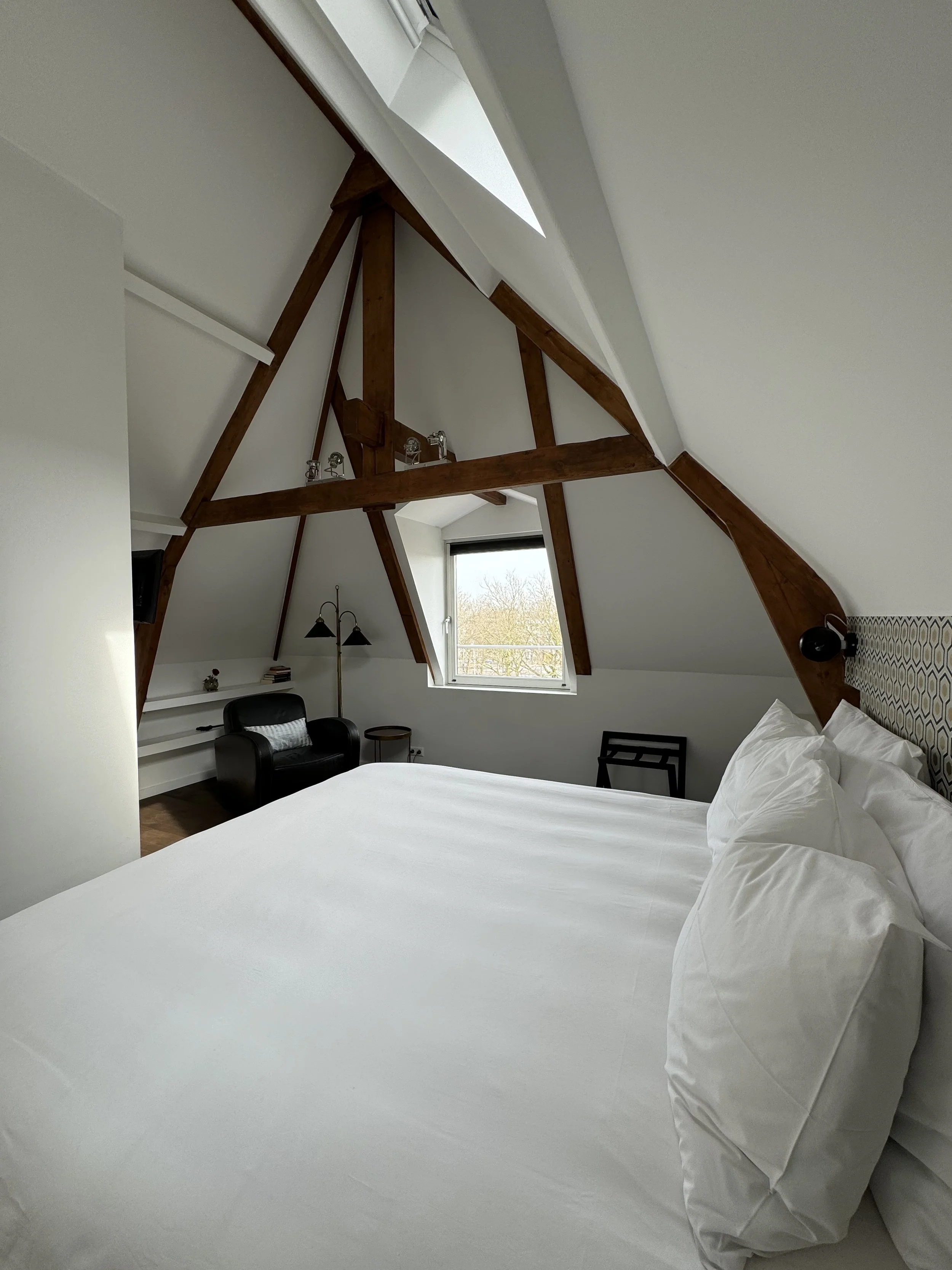 A bedroom with sloped ceilings and exposed wooden beams, featuring a large bed with white linens, a black armchair, a small side table, and a window showing trees outside.
