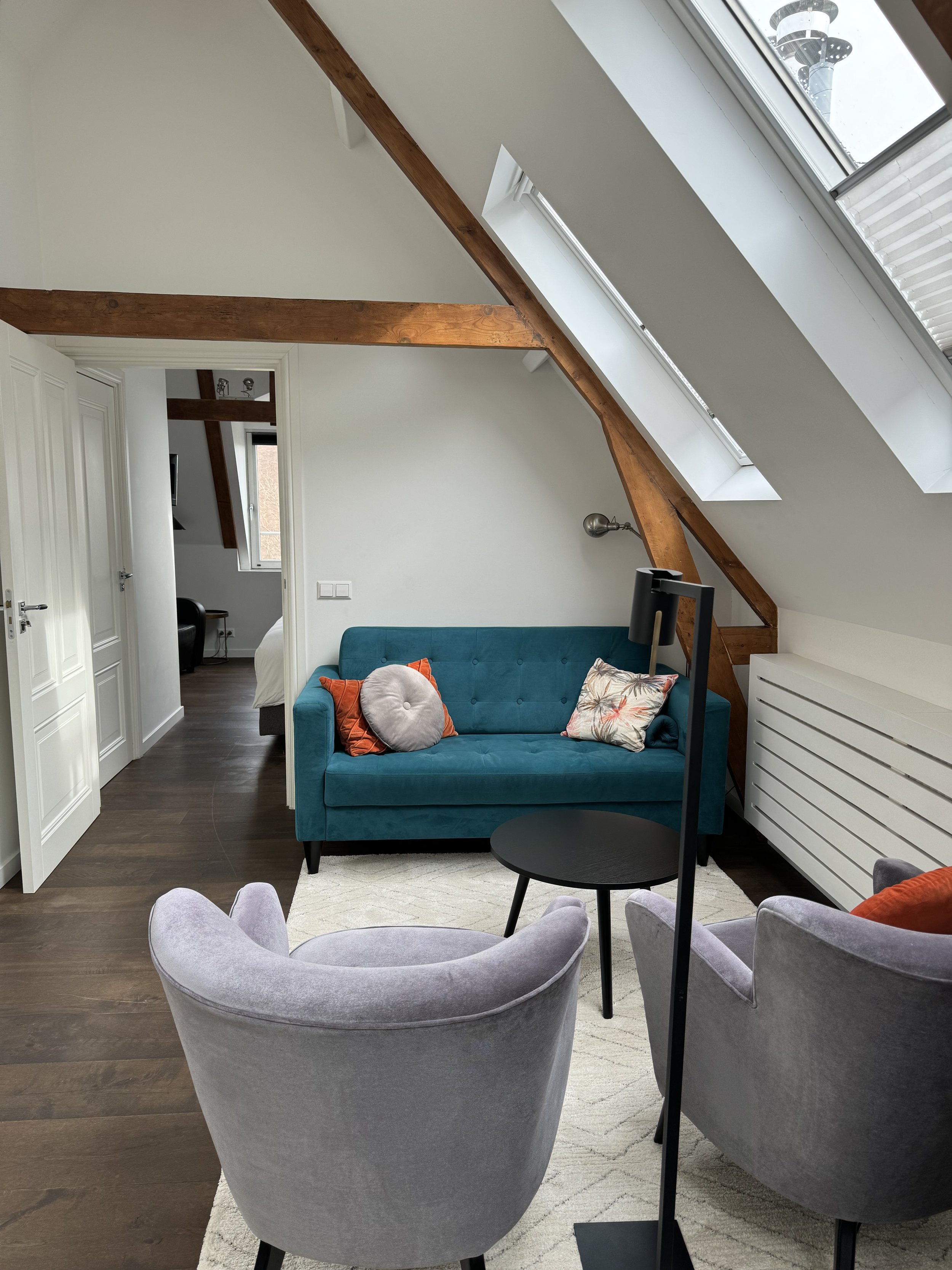 A cozy living room with a teal sofa, a round gray velvet armchair, and multiple pillows, set against sloped ceilings with wooden beams and skylights.