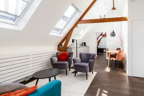 Living room and dining area with sloped ceiling, skylights, gray chairs, a small black coffee table, a wooden dining table, and pink and orange chairs.