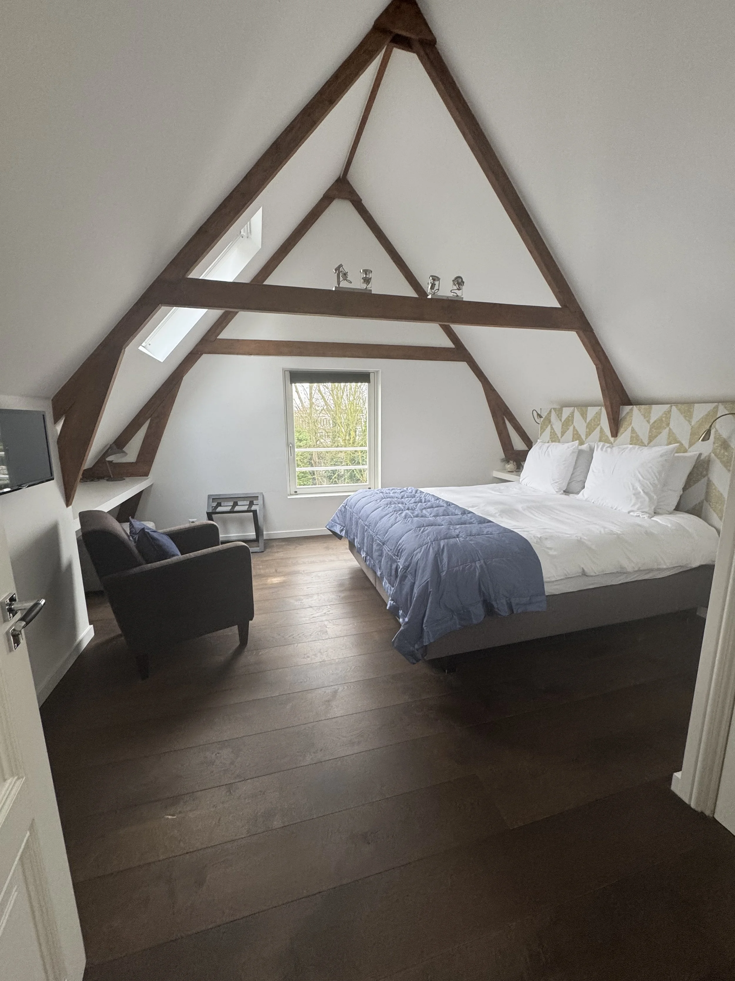 A bedroom with sloped ceilings and exposed wooden beams, a bed with white pillows and a blue comforter, a window with a view of trees, a black armchair, a small side table, and a wall-mounted TV.
