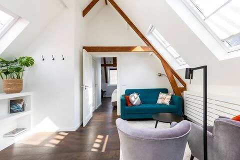 Cozy attic living space with blue sofa, armchair, small black table, white walls, skylight windows, and wooden beams.
