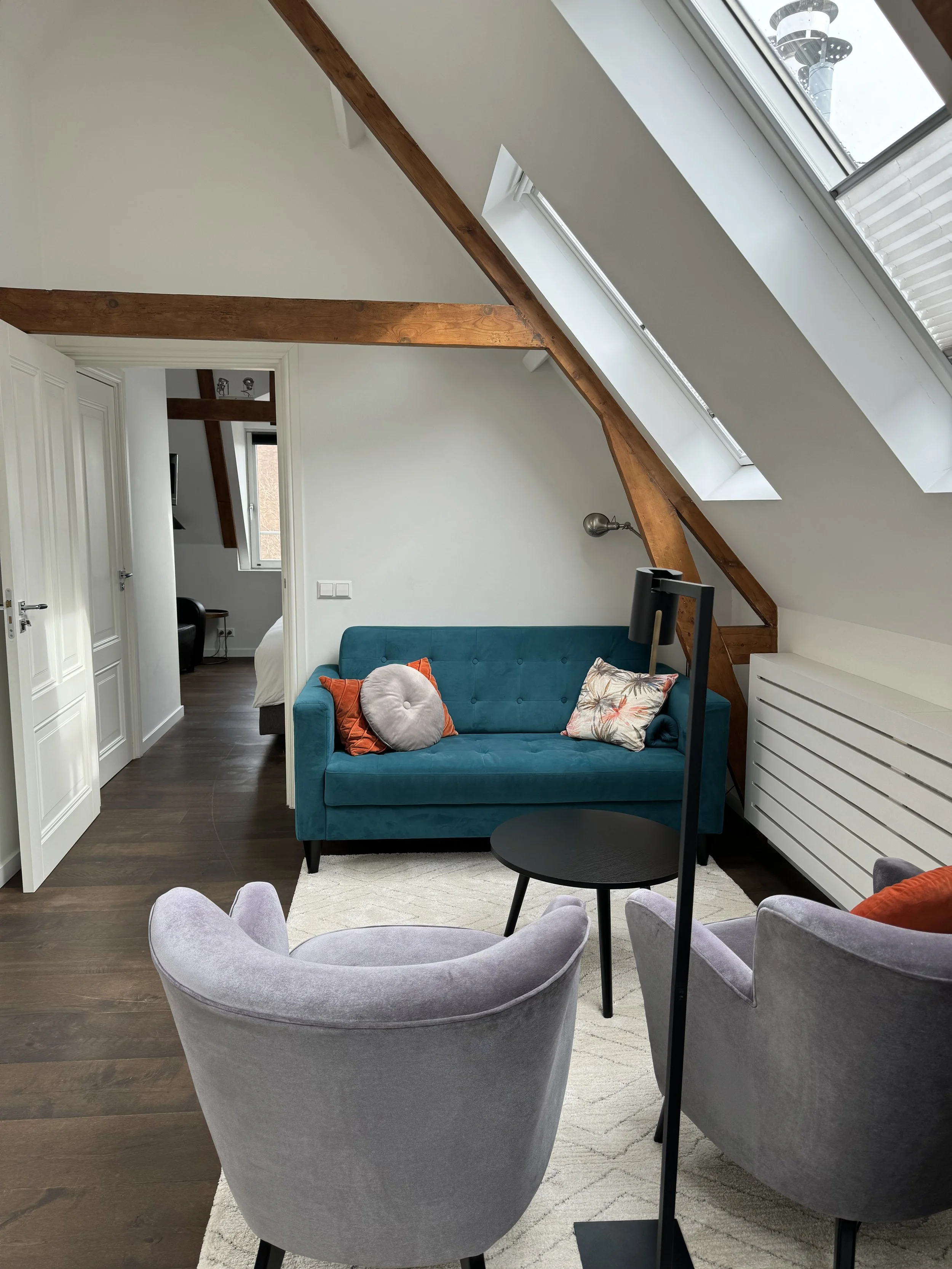 Living room with a teal sofa, two gray armchairs, a black side table, a white area rug, and wooden beams. Skylight windows on the sloped ceiling.