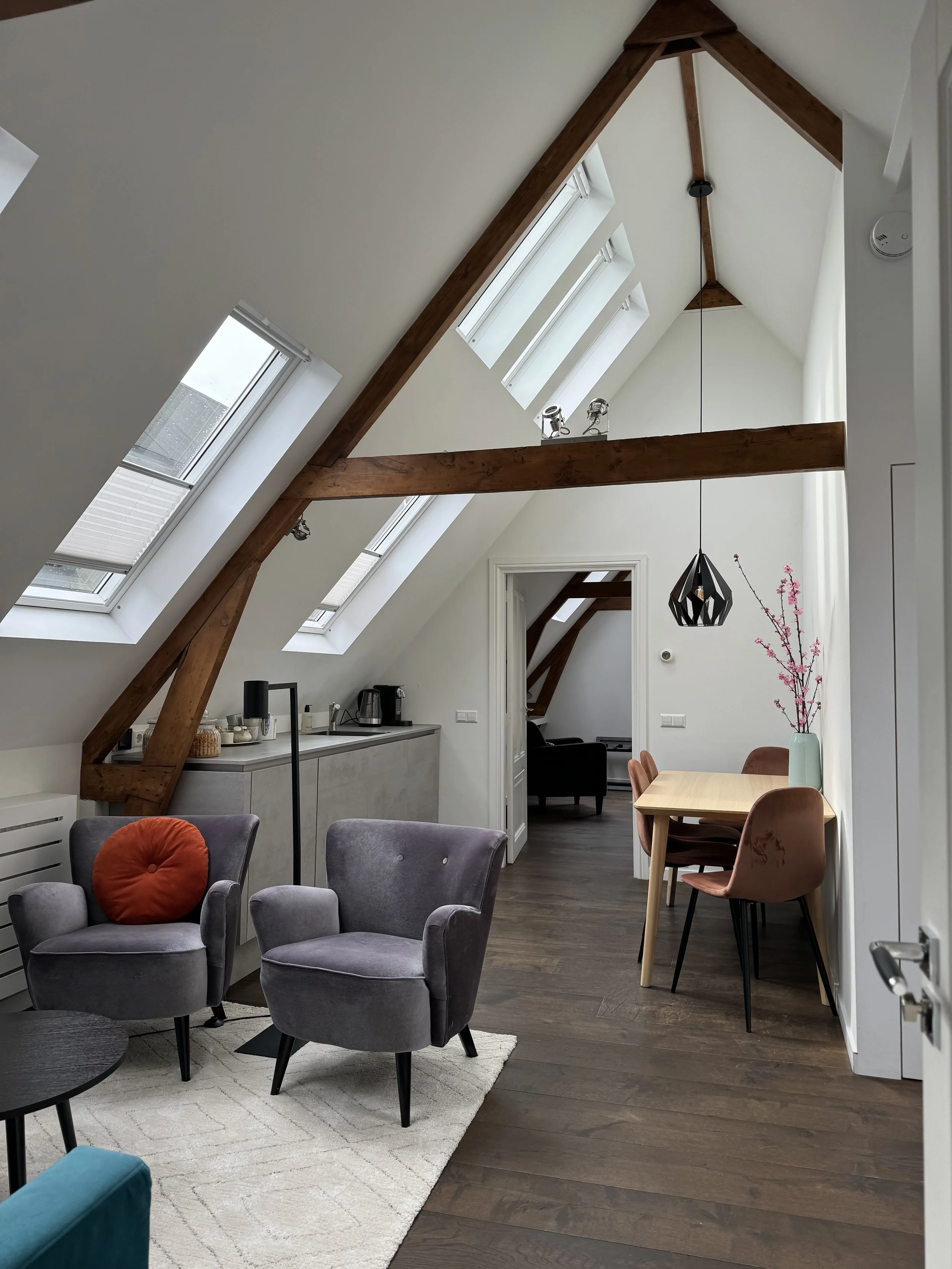 Interior of an attic apartment with sloped ceilings and skylights, featuring a sitting area with two purple armchairs, a round black side table, a dining table with four chairs, a vase with pink flowers, and a kitchen section with appliances.