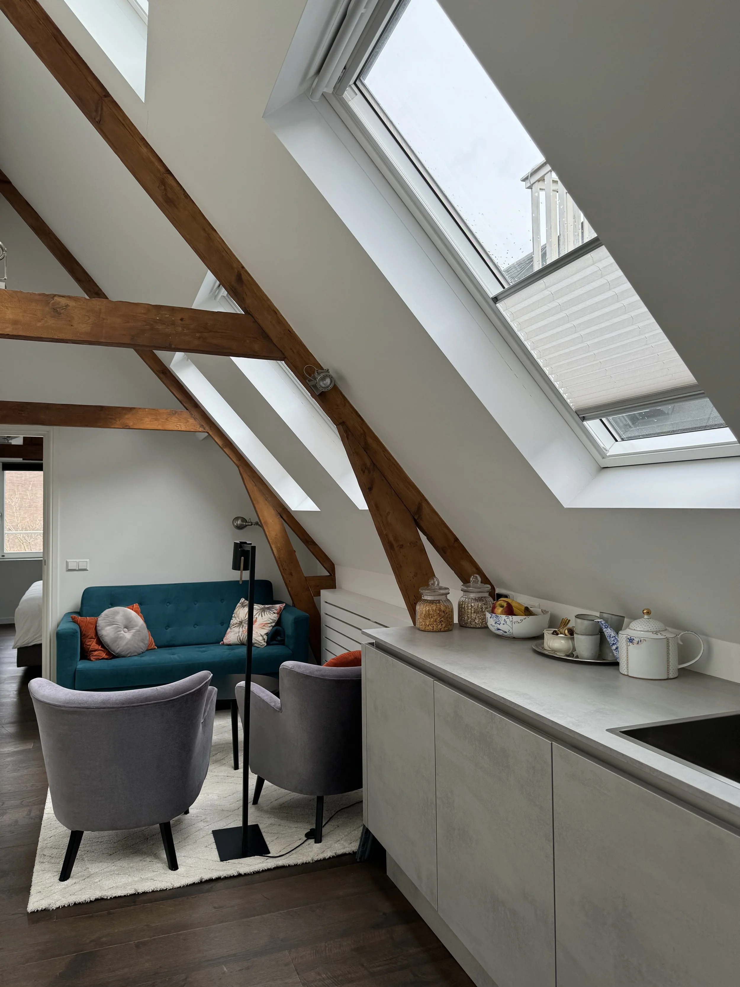 Cozy attic living space with a sloped ceiling and large skylight window, featuring a teal sofa, two gray armchairs, and a small white rug, with a kitchen counter and decorative jars on the side.