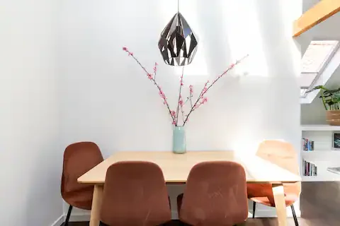 A dining table with a decorative vase holding pink flowers, surrounded by four brown chairs, under a modern black pendant light, with a white wall and bookshelf in the background.