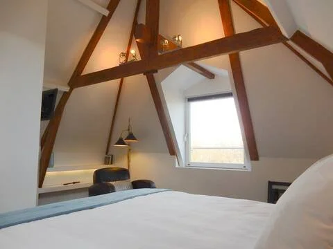 Bedroom with a slanted ceiling, exposed wooden beams, a window with a view outside, a bed with white sheets, and a desk with a lamp.