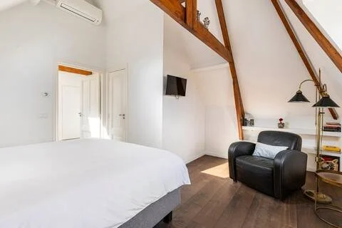 A bedroom with a white bed, black armchair, wall-mounted TV, and a small bookshelf with decor, wooden beams on the ceiling, and hardwood flooring.
