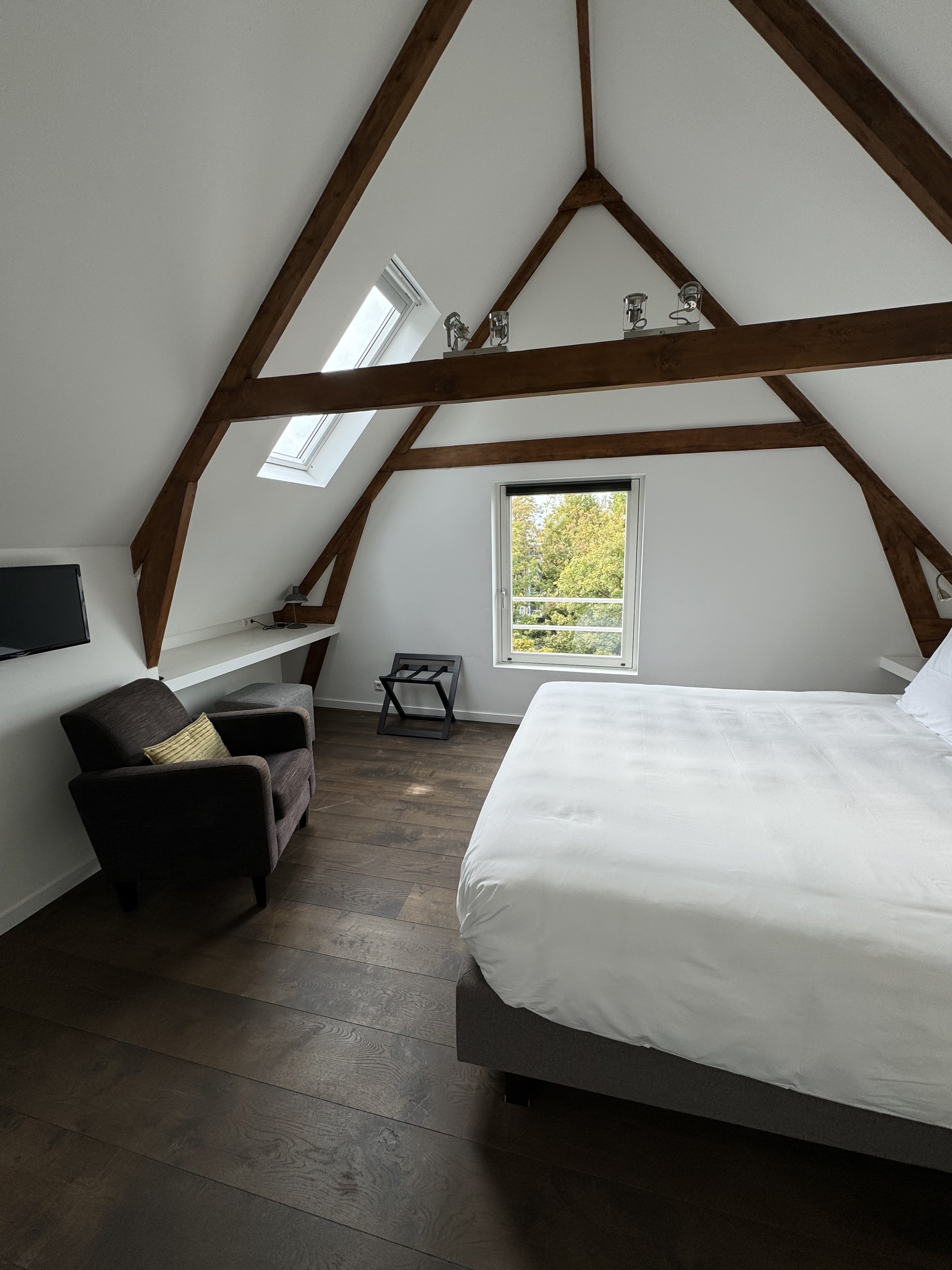 A bedroom with a sloped ceiling featuring exposed wooden beams, a large bed with white bedding, a small armchair, a window, and a desk area with a chair.