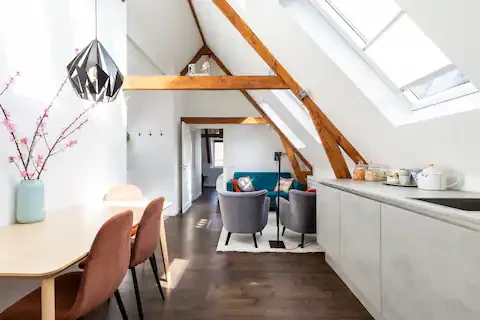 Living room with sloped ceiling, skylight windows, and modern furniture including a dining table with chairs, a vases with pink flowers, a seating area with armchairs, and a white kitchen counter.