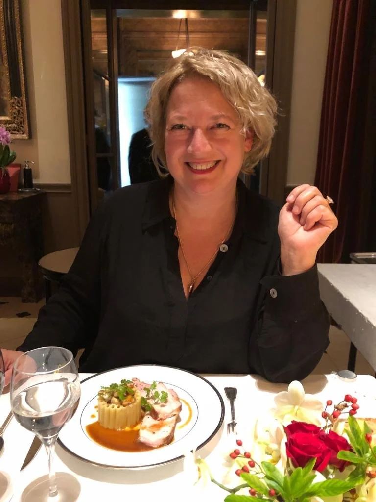 A woman with short blonde curly hair smiling and sitting at a dinner table with a plated dish in front of her, surrounded by flowers, a glass of water, and silverware, in a warmly lit restaurant.
