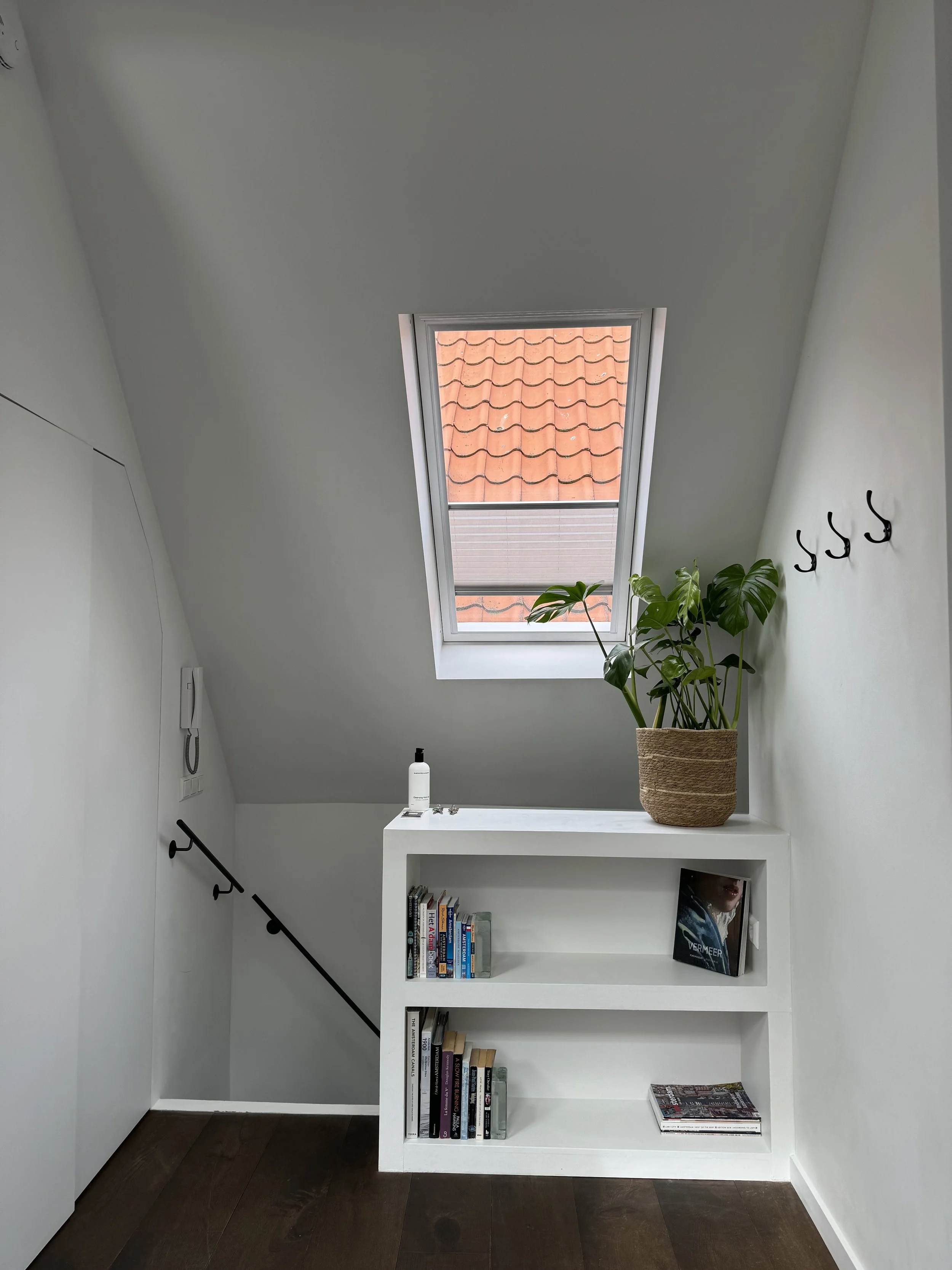 A small white bookshelf with three shelves, a potted plant on top, and a window with a red tiled roof visible outside. Walls are white, and there are black hooks and a black handrail.