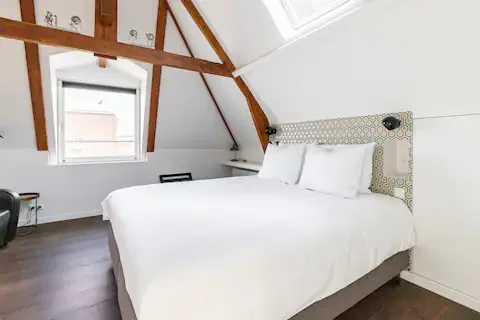 A neatly made white bed with pillows in a bright attic bedroom with wooden beams and a skylight window.