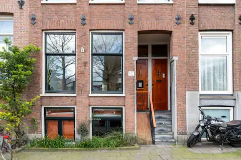 Facade of a brick apartment building with a wooden front door, steps, and two large windows. Bicycles and a motorcycle are parked outside.