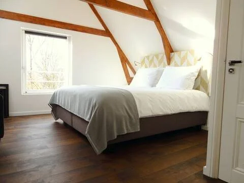 A neatly made bed with white pillows and yellow patterned pillows in a cozy attic bedroom with wooden beams and a window.