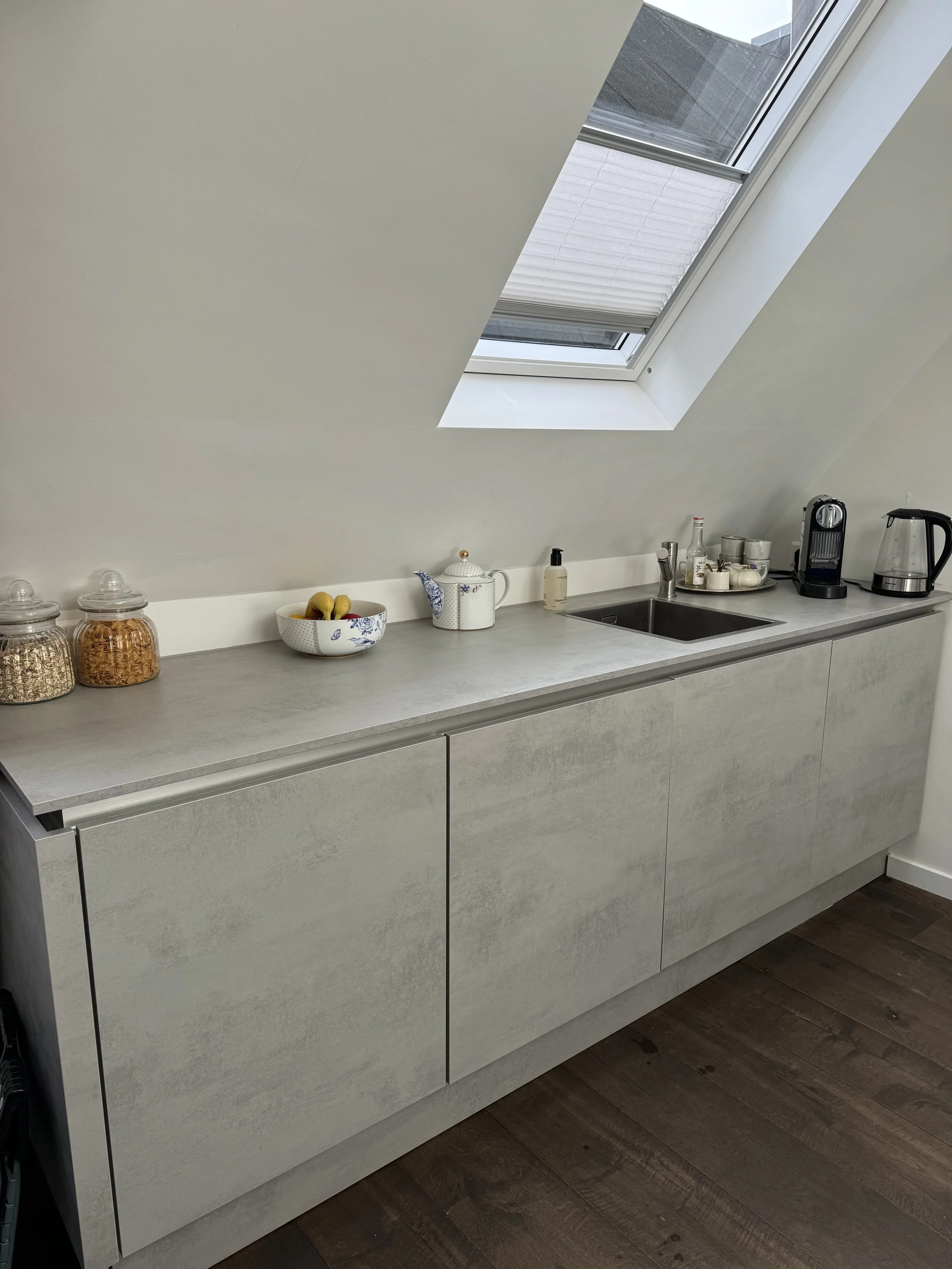 Modern kitchen with a sloped ceiling and a skylight window, featuring a countertop with jars, a bowl of fruit, a teapot, soap dispenser, and coffee machine.