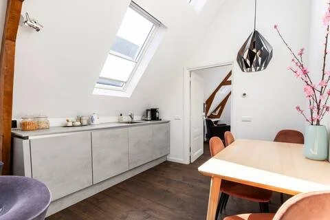 Modern dining area with a wooden table, four chairs, a large vase with pink blossoms, and a black geometric pendant light, in an attic space with a sloped ceiling and a skylight.