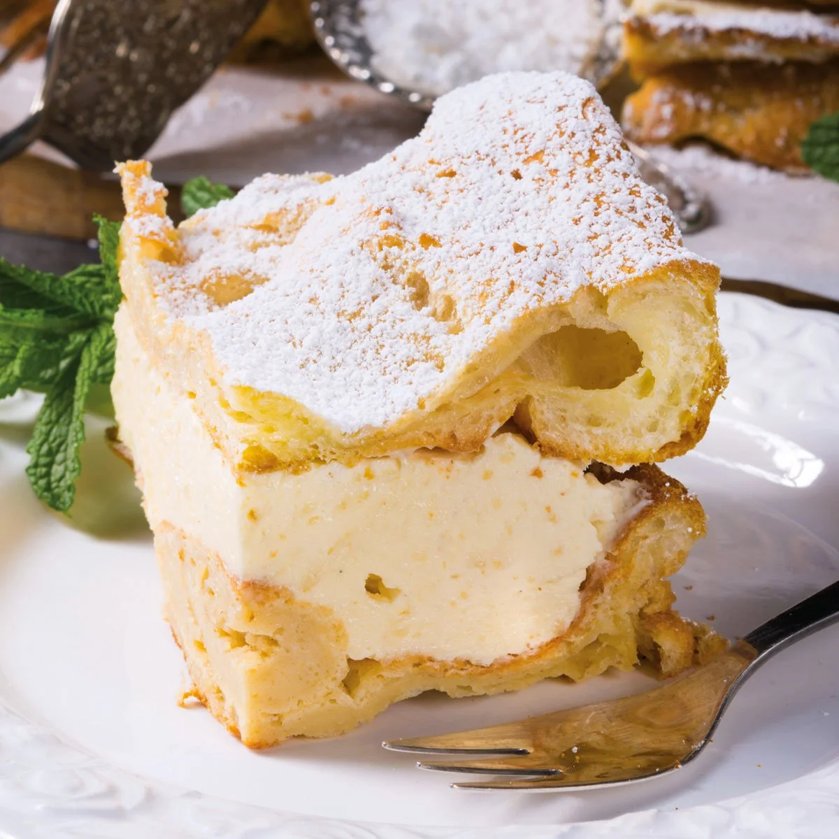 A slice of layered lemon shortcake with whipped cream and powdered sugar, garnished with mint leaves, on a white plate.