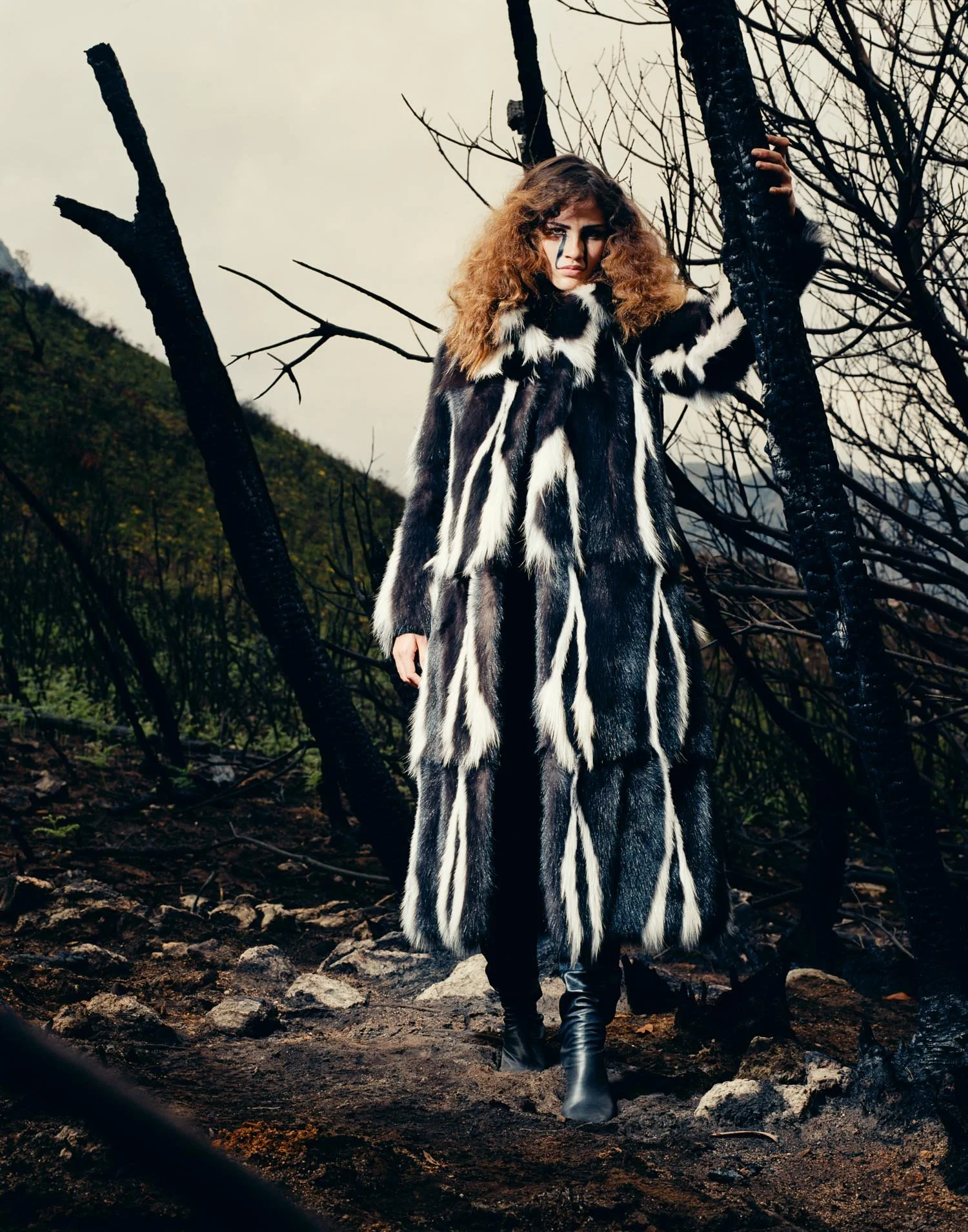 A woman in a long, black and white fur coat standing outdoors in a desolate landscape with burnt trees and dark soil, under an overcast sky.