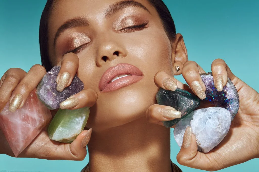 Woman with makeup posing with large, colorful geodes and crystals against a bright blue background.