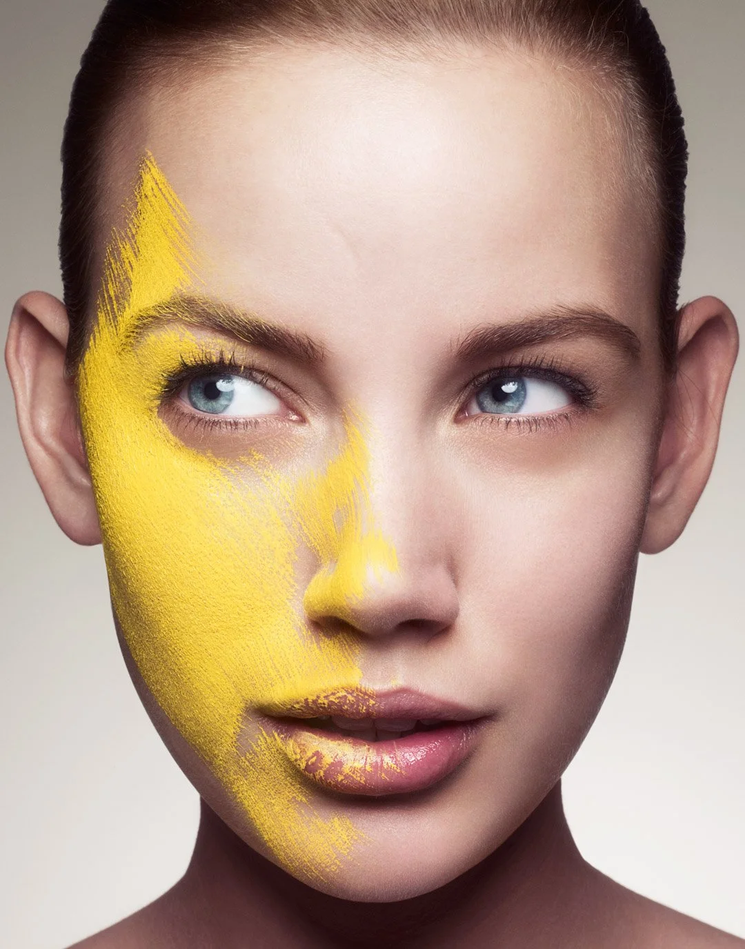 Close-up of a woman with blue eyes and fair skin, with yellow paint on the left side of her face.