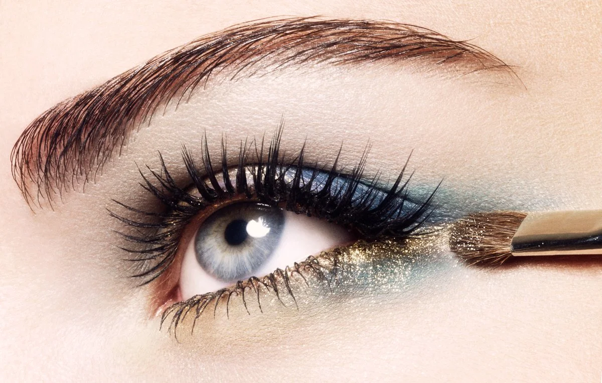 Close-up of a person's eye with makeup; gold glitter eyeshadow on the lower lid, blue eyeshadow on the upper lid, and black mascara on the eyelashes. A makeup brush is applying eyeshadow to the lower lash line.