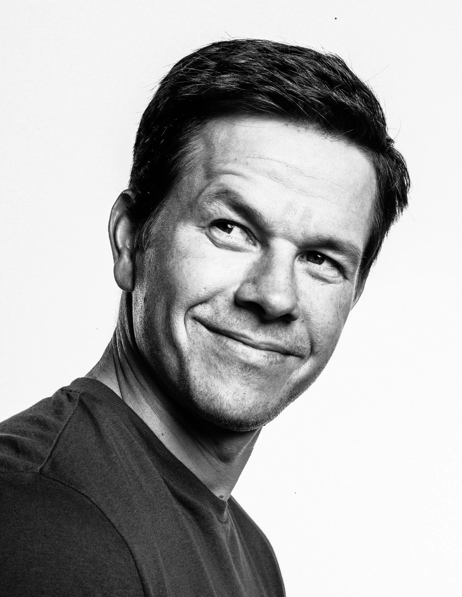 Black and white portrait of MARK WAHLBERG with short dark hair, smiling slightly, wearing a dark shirt, against a plain light background.