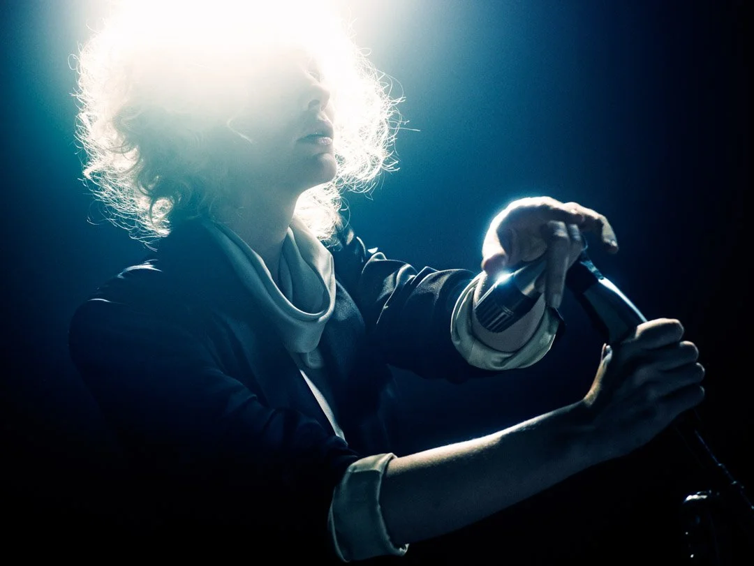 CATE BLANCHETT with curly blonde hair holding a microphone on stage with bright backlighting.