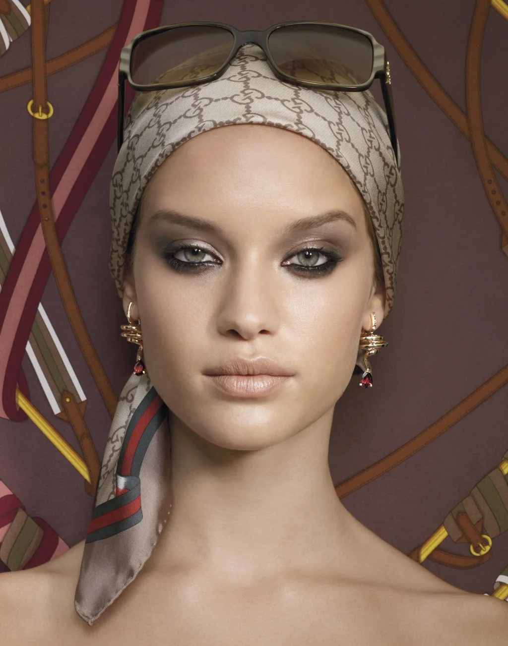 A woman wearing a beige patterned headscarf with sunglasses on top, gold earrings, and makeup, posed against a backdrop of leather straps and belts.