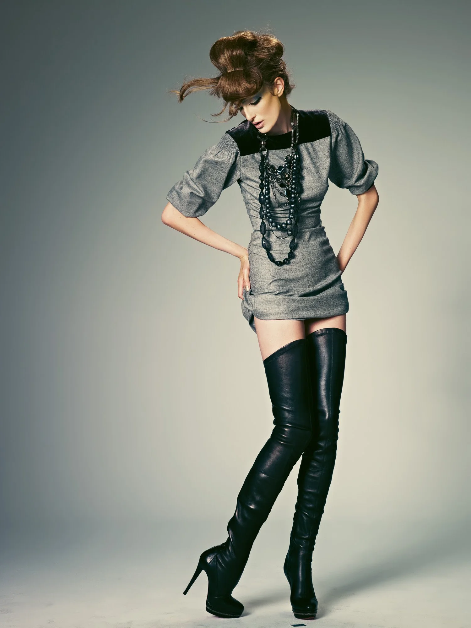 A woman with long, wavy hair styled in an updo poses with her head bent down, wearing a gray and black dress with puffed sleeves, layered black necklaces, and over-the-knee black leather high-heeled boots.